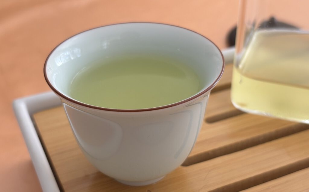Eine weiße Teetasse mit hellgrünem Sencha Tee steht auf einem Holztablett. Rechts daneben ist eine teilweise sichtbare Glaskanne mit Tee.