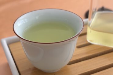 Eine weiße Teetasse mit hellgrünem Sencha Tee steht auf einem Holztablett. Rechts daneben ist eine teilweise sichtbare Glaskanne mit Tee.